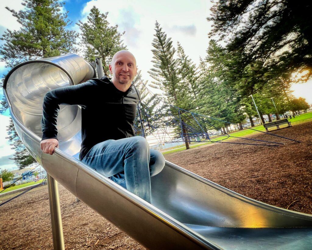 Photo of Dave squatting on a slide in a playground, with Norfolk Island pine trees and the sunset in the background.